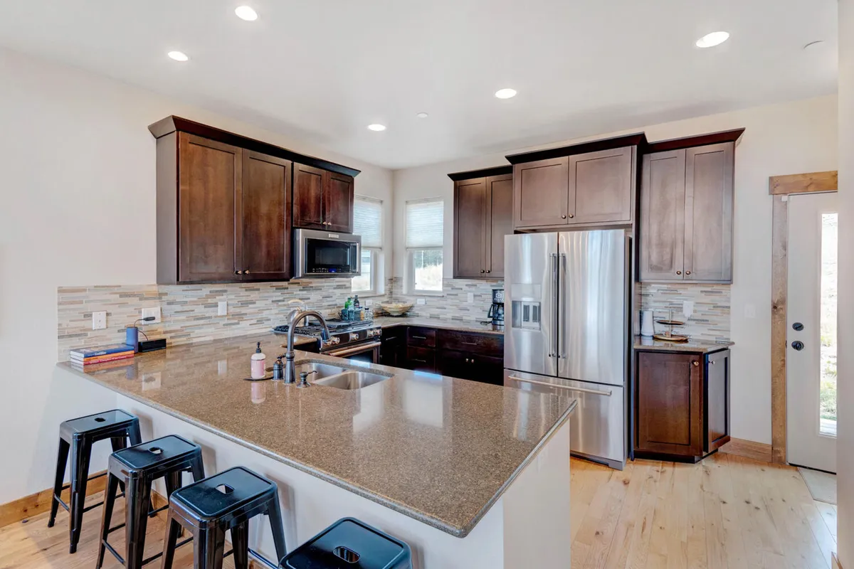 A spacious, open-concept kitchen featuring a large granite countertop, dark wooden cabinets, stainless steel appliances, and several bar stools. The space is well-lit and designed for both meal preparation and social interaction.