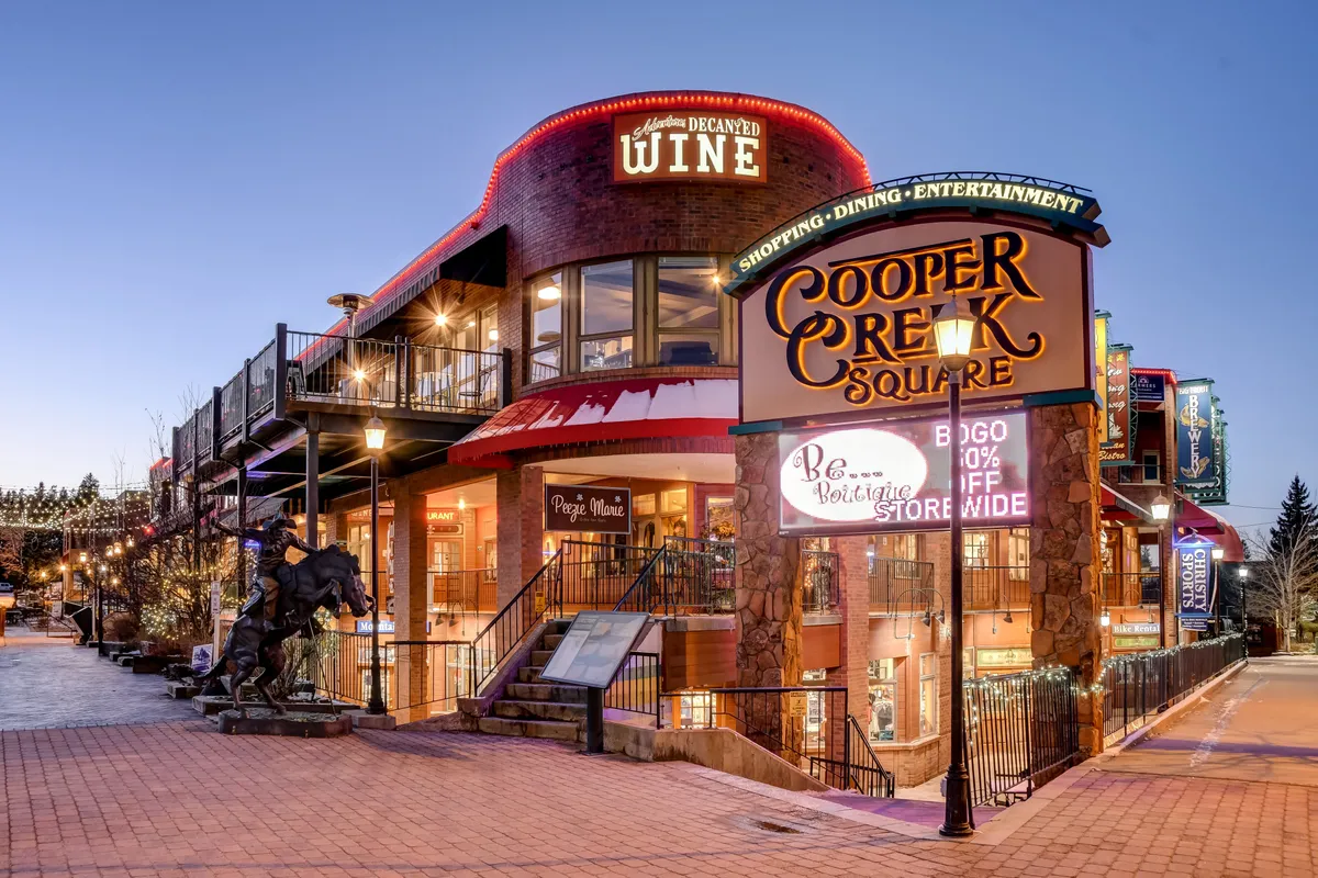 A lively scene at Cooper Creek Square featuring a prominent sign, wine shop signage, staircase to upper levels, and a horse sculpture. People can be seen enjoying the festive atmosphere, surrounded by boutique shops and hints of vibrant local activity.