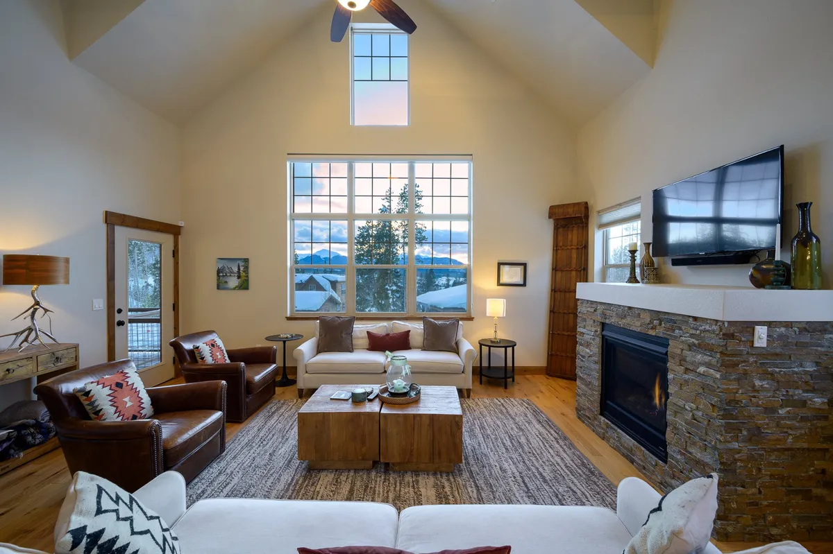 A spacious main living area featuring a soaring vaulted ceiling, large windows showcasing panoramic mountain views, a striking stone fireplace, a plush seating arrangement, and a wooden coffee table. Soft natural light pours in, inviting guests to relax, read, socialize, or simply soak in the scenery.