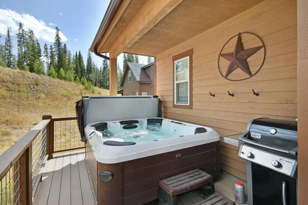 This image features a wooden deck surrounded by trees, equipped with a private hot tub and a gas grill. The setting is tranquil and inviting, perfect for outdoor relaxation and social gatherings. The deck is spacious enough to host group activities, and the natural landscape enhances the sense of retreat.