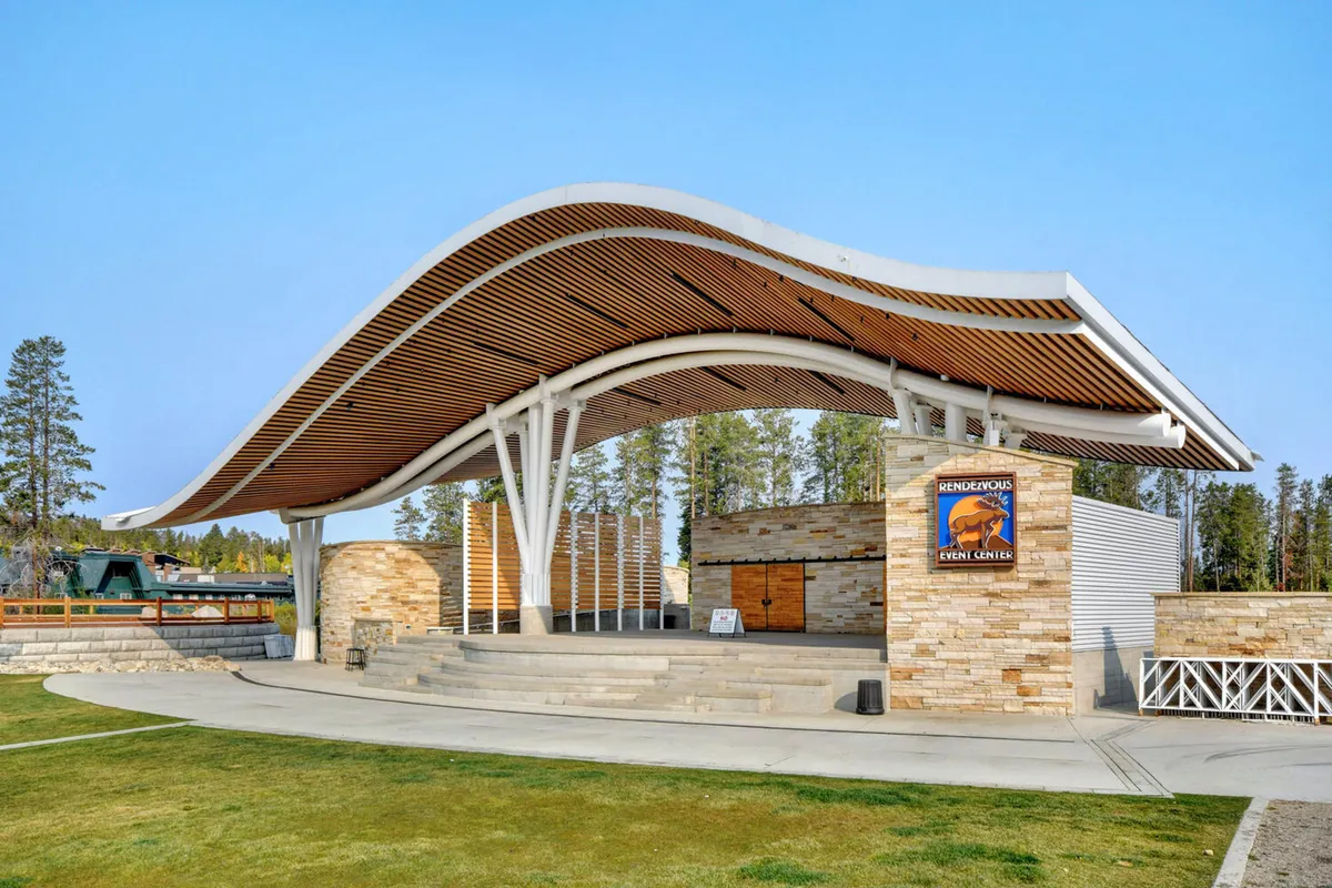 The outdoor Hideaway Park amphitheater, with its distinctive curved roof structure and stone wall, is set up for an event. Event signage and an open plaza hint at upcoming concerts or community gatherings, inviting visitors to join in the festivities.