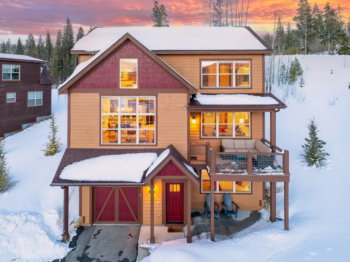 The image showcases the front exterior of a luxurious single-family mountain villa in Fraser, Colorado. The scene features a welcoming front entrance, expansive windows revealing warm indoor lighting, and a spacious deck perfect for gathering. Snow blankets the yard, inviting guests to play or relax while enjoying breathtaking mountain views at sunset.