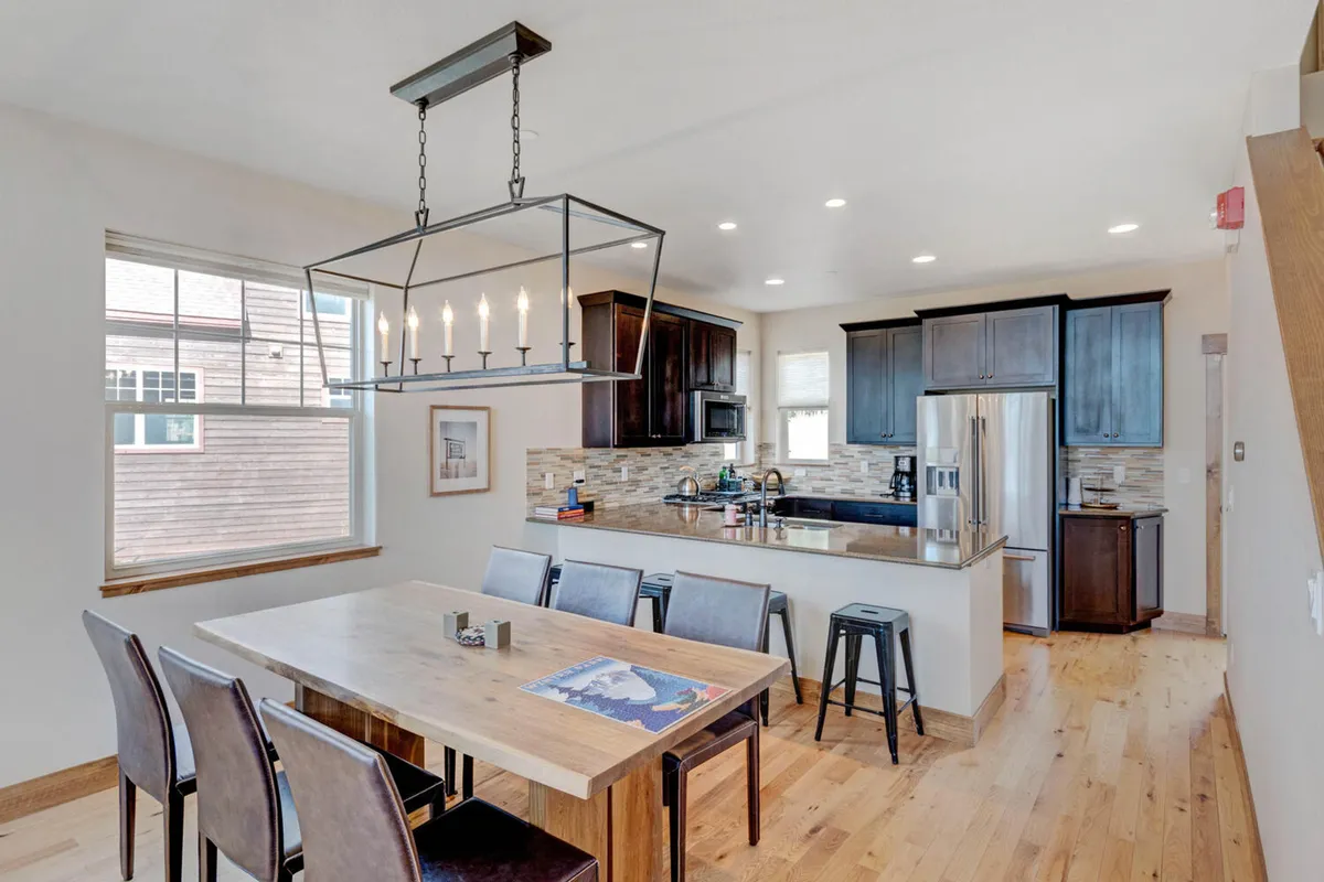 A warm and welcoming dining area with a large dining table set beneath a chandelier, adjacent to a kitchen island. The space offers a scenic window view, suggesting a cozy setting for family meals or entertaining guests.