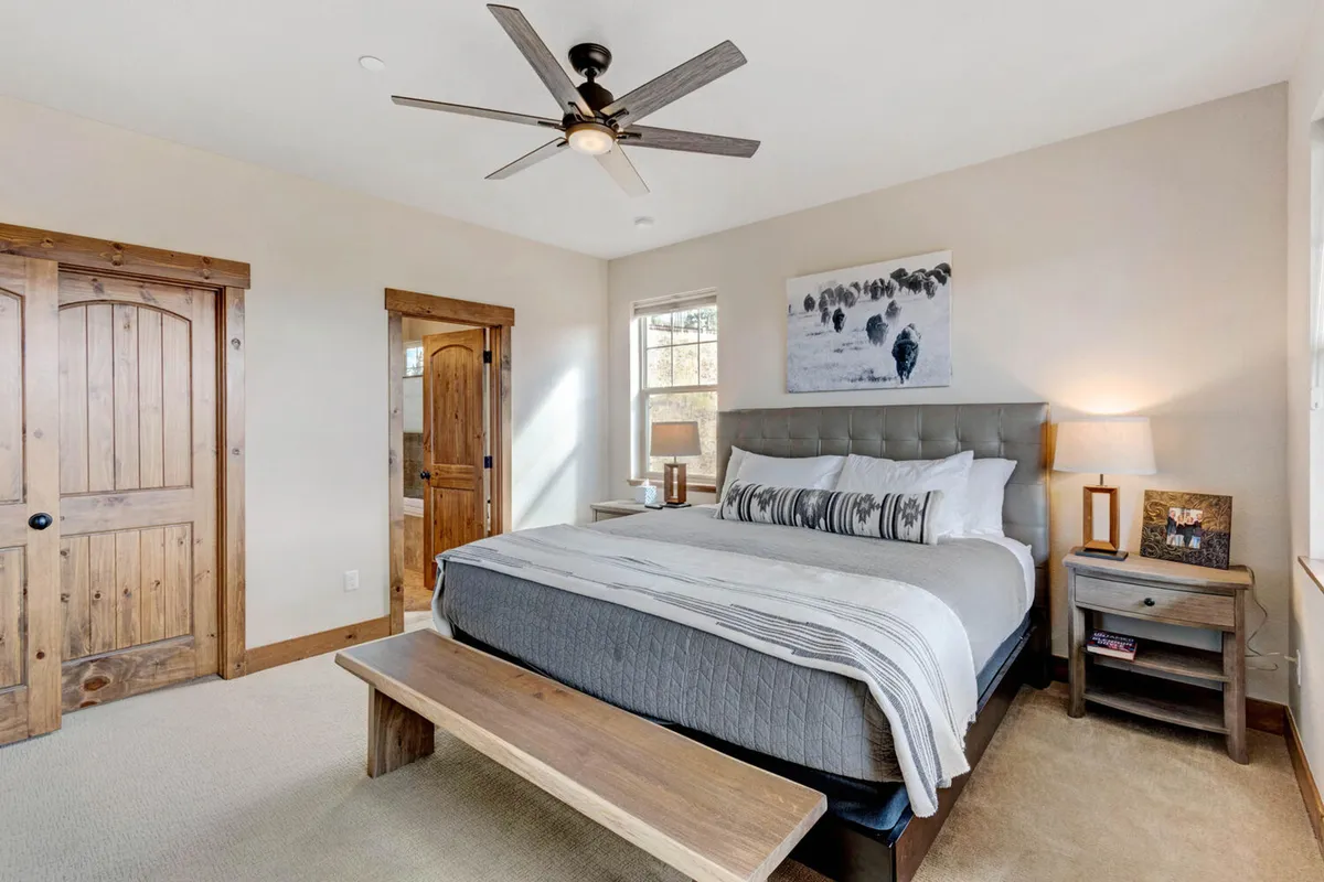 A tranquil bedroom featuring a plush bed with premium linens, wooden doors, and soft lighting from a nightstand lamp. Artwork above the bed and sunlight streaming in create a serene, restful atmosphere ideal for unwinding.
