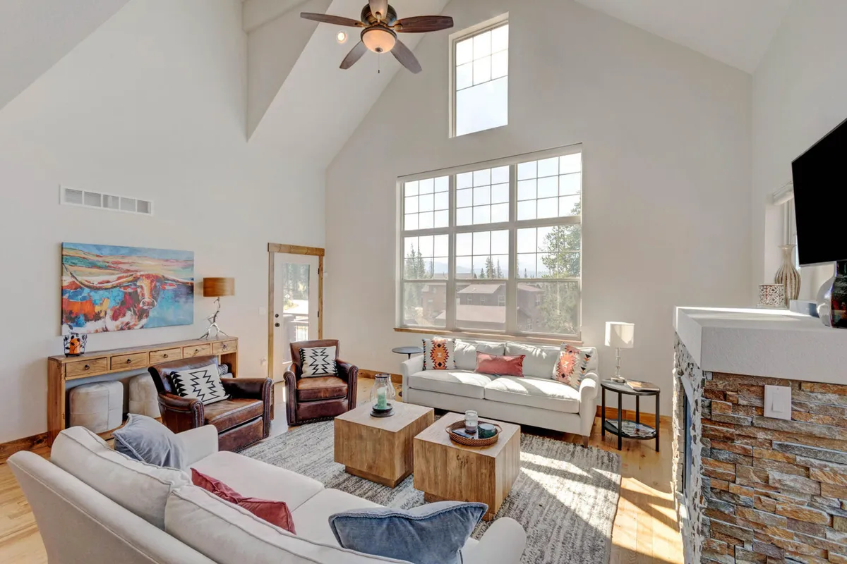 An inviting living room with a comfortable seating group, textured stone fireplace, large windows flooding the space with light, and tasteful mountain-inspired artwork. The layout is ideal for socializing, enjoying family game nights, or unwinding with a book.