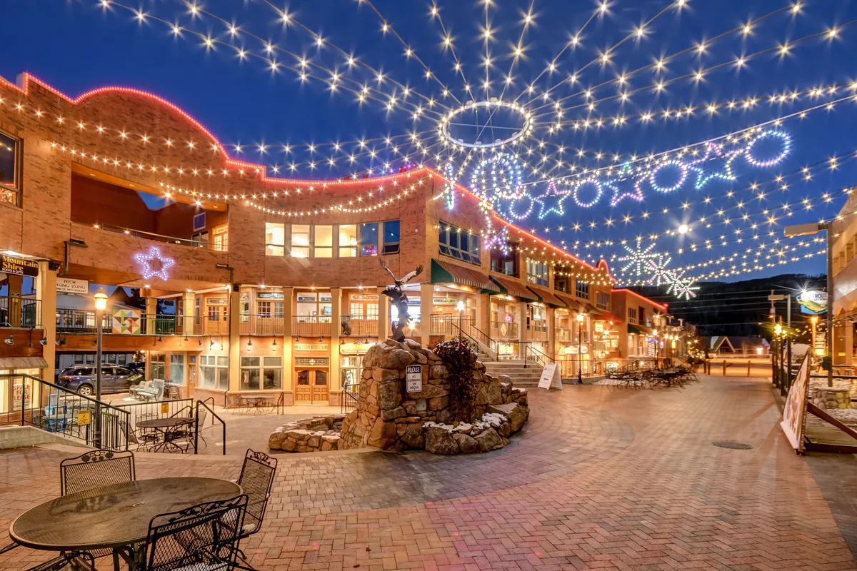A beautifully lit outdoor plaza at night, strung with twinkling lights and decorative stars around a central fountain. Storefronts and people strolling create an inviting, festive mood perfect for evening gatherings.